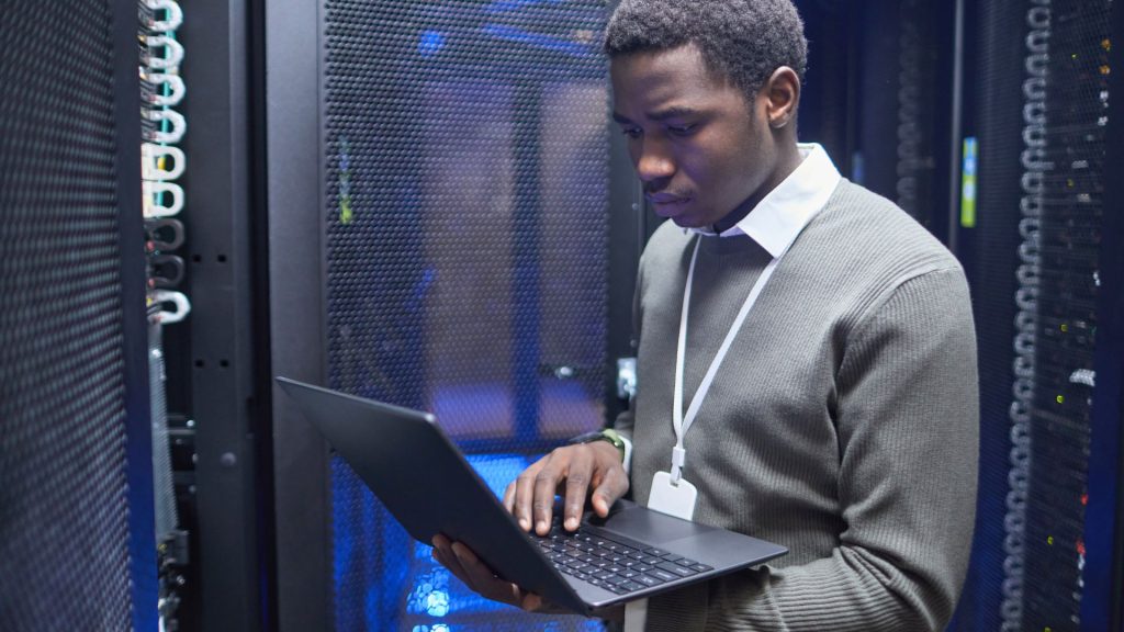 A man in a server room holds a laptop, surrounded by rows of servers and blinking network lights.