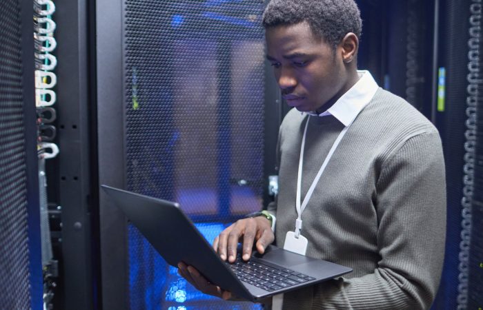 A man in a server room holds a laptop, surrounded by rows of servers and blinking network lights.